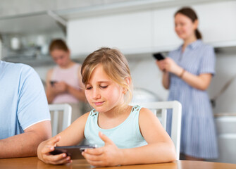 Preschooler girl sitting near kitchen table with phone in hands and playing online game, mom and older sister surf Internet in background, unfocused