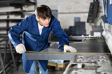 Young guy worker measures sheet of metal with tape measure in metallurgical workshop
