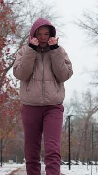 Young woman dressed in warm winter clothing walks through a snowy park, holding her hands close to her face for warmth, bare trees, red berries, and distant figures
