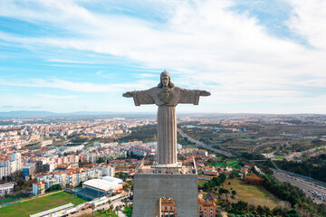 Drone shot of Sanctuary of Christ the King in Almada, Portugal. Iconic statue with open arms overlooks city buildings under blue sky. Famous landmark and tourist attraction