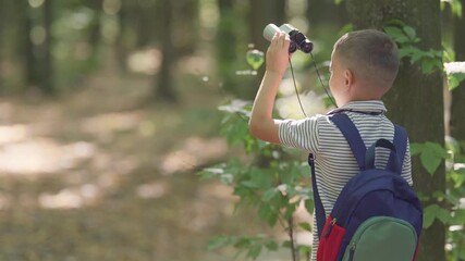 A young boy with a backpack uses binoculars to observe the surroundings. He is exploring the outdoors on a sunny day in a wooded area.