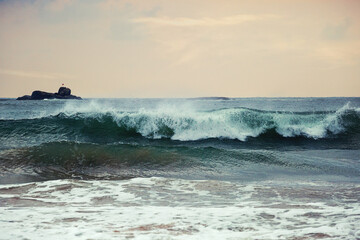 Powerful ocean wave crashing near rocky island at sunset