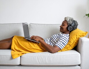 Relaxed African American woman lying on sofa, using laptop and headphones for work or leisure. Enjoying comfort and connectivity at home.