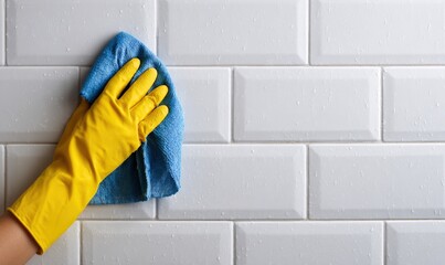 A hand in a yellow glove wipes a blue cloth across a white tiled wall