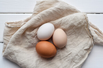 Top down view of three different chicken eggs — white, brown, and beige — resting on a linen...