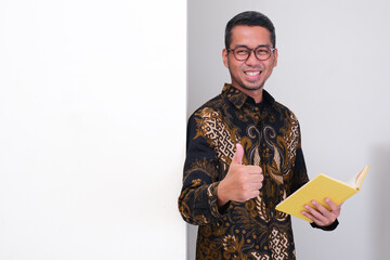 Indonesia school teacher standing beside a wall holding book and give thumbs up with happy face