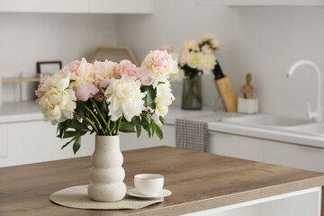 Vase with bouquet of beautiful peonies and cup of tea on table in kitchen, closeup