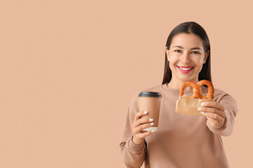 Young happy woman with tasty pretzel and cup of coffee on beige background