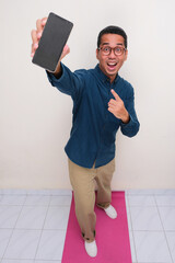 High angle view of adult asian man standing while showing blank phone display screen with amazed face