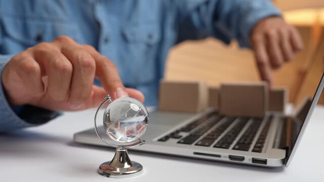 A hand spins a globe while the other arranges e-commerce packages on a laptop, symbolizing global trade, online shopping, worldwide logistics, digital entrepreneurship, and cross-border commerce.
