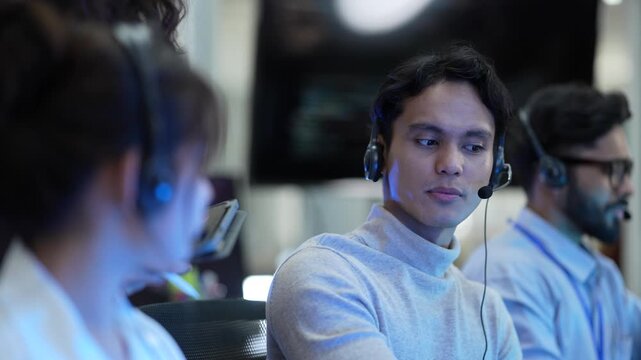 A friendly team of operators with headsets smiles while talking to customers. The diverse group works in a busy call center, helping people and providing good service