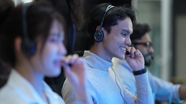 A friendly team of operators with headsets smiles while talking to customers. The diverse group works in a busy call center, helping people and providing good service.