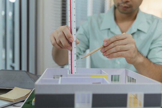 Asian male architect sitting at a desk, measuring a modern house model with triangular ruler and pencil. Office setup includes design tools, blueprints, and safety gear, suggesting architectural work.