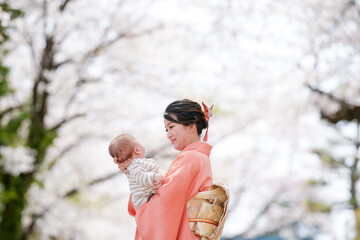 A woman is holding a baby in her arms while wearing a pink kimono. The scene is set in a park with...