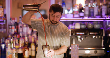 Cocktail making. Shaking drink. Professional mixologist working in pub with neon light. Male bartender pouring pink strained liquid.