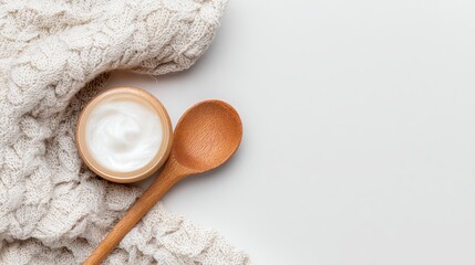 Creamy white substance in a wooden bowl near a wooden spoon atop a folded, textured beige knit fabric against a blank, off-white background