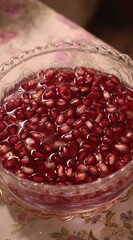 A glass bowl filled with bright red pomegranate seeds submerged in water, displayed on a floral tablecloth, lit warmly