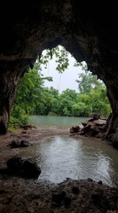 View from a dark cave entrance showing a body of water surrounded by green trees and foliage. Rocks and dirt in the cave