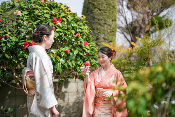 Two women in traditional Japanese clothing are standing in a garden, one holding a red flower....