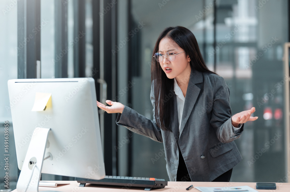 Poster asian manager sitting at desk, staring at computer screen with a confused expression, raising hands 