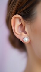 Macro shot of a woman’s ear with classic round pearl stud earrings, soft blurred pastel background for elegance.
