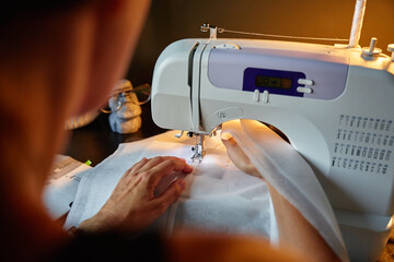 Close up of woman using modern sewing machine to stitch fabric at home under lamp light. Concept of sewing and tailoring