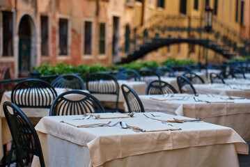 Empty outdoor restaurant tables with tablecloths and cutlery in Venice, Italy. Street cafe with bridge over Venetian canal on background in morning. Concept of dining, tourism and travel