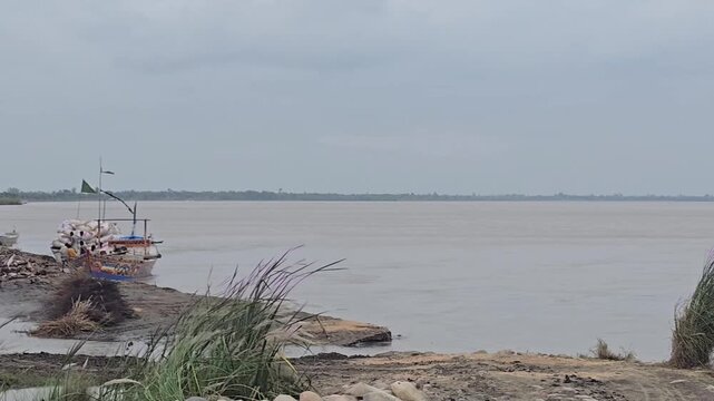 Rustic fishing boat on a wide muddy river, traditional wooden vessel docked at a rural riverbank, overcast tropical landscape with murky water