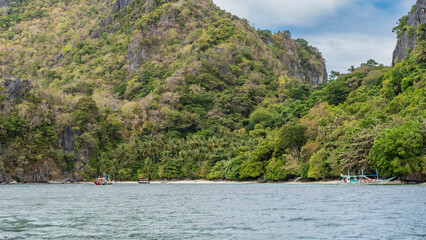 The beautiful tropical beach is visible from the ocean. White sand, thickets of palm trees, green bushes. The boats near the shore. Mountains, blue sky, clouds. Turquoise sea in the foreground. 