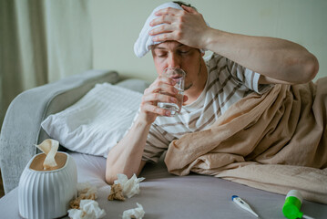 Sick man with a wet towel on his forehead is lying in bed covered with a blanket, drinking water, with used tissues, a thermometer and a nasal spray on the mattress