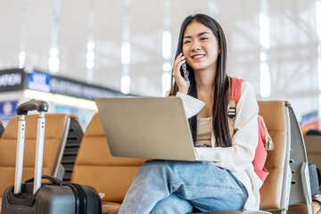 Smiling young asian woman sitting in waiting room and talking over smartphone with friend or family and using laptop. Beautiful female traveller talking on cell phone at airport lounge.