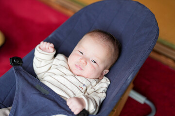 A baby is laying on a blue high chair. The baby is wearing a striped shirt and has a serious expression on its face