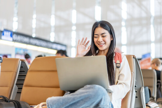Attractive asian young woman traveler talking on video call to friend or family through computer laptop while waiting for her flight sitting on seat in departure gate at airport terminal.