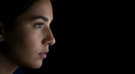 A serious young woman is captured in a dramatic low-key close-up profile shot, gazing intently forward with intense mood against a solid black background.
