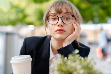 Professional woman with glasses rests her chin on her hand while enjoying coffee at an outdoor cafe...