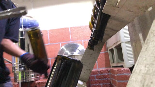 Asian man scalds a wine bottle in boiling water to pasteurize it, then places it on a wooden rack while  working in a small local winery.