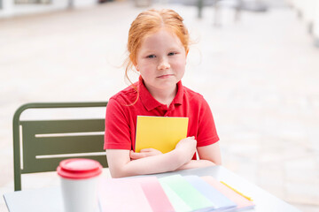 Young girl with red hair sitting at a table in a sunny outdoor setting, holding a yellow notebook...