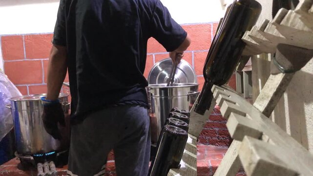 Asian man scalds a wine bottle in boiling water to pasteurize it, then places it on a wooden rack while  working in a small local winery.
