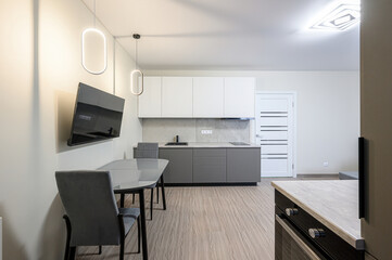 Modern kitchen with gray cabinets, a dining table, and a wall-mounted TV. The space is bright and minimalist with pendant lights