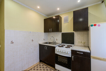 compact kitchen with dark wood cabinets, a white stove, and a stainless steel sink. The walls are tiled, and the upper part painted a pale yellow. A white refrigerator is seen on the right