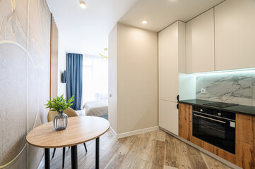 modern studio apartment featuring a dining area with an oval table, a glimpse of a bedroom, and a compact kitchen with white cabinets and wood accents