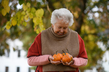 Portrait of elderly woman on walk in the park during warm autumn day, holding handful of pumpkins.