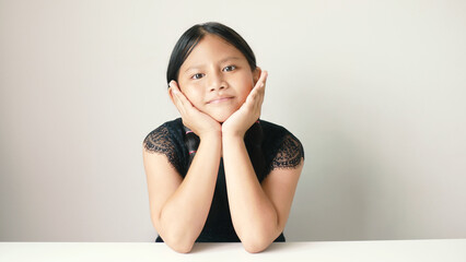 A small Asian girl sits with a sweet smile, gently supporting her chin with both hands on a white...