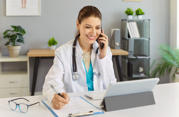 Cheerful young female doctor or nurse sitting at her workplace at the desk in office talking by...