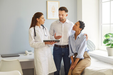 Fototapeta premium Consulting in clinic, female reproductologist doctor speaking holding tablet, helping couple, husband supporting wife listening information, professional and caring serious discussion, medical care