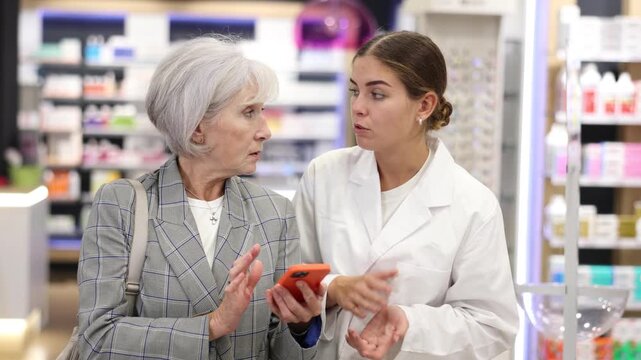 Concerned old female customer asking young chemist about product reflected on her phone in large pharmacy. High quality 4k footage