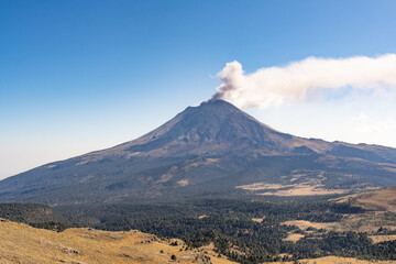 A mountain with a volcano on top and a cloud of smoke rising from it. The sky is clear and blue. Iztacihuatl popocatepetl volcano mexico, © Alejandro