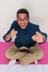 High angle view of adult asian man sitting cross legged pointing at the camera with excited face