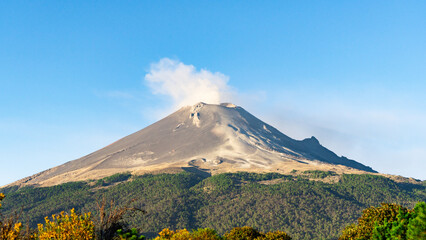 Fototapeta premium A mountain with a cloud of smoke rising from it. The sky is clear and blue. The mountain is covered in trees and he is covered in snow. Iztacihuatl popocatepetl volcano mexico,