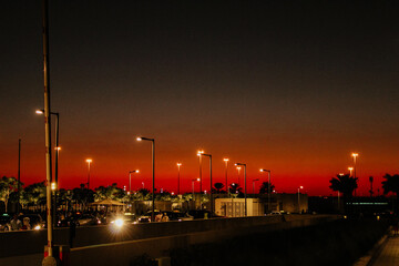 A vivid red and orange sunset casts an intense glow over an urban parking lot, with streetlights,...